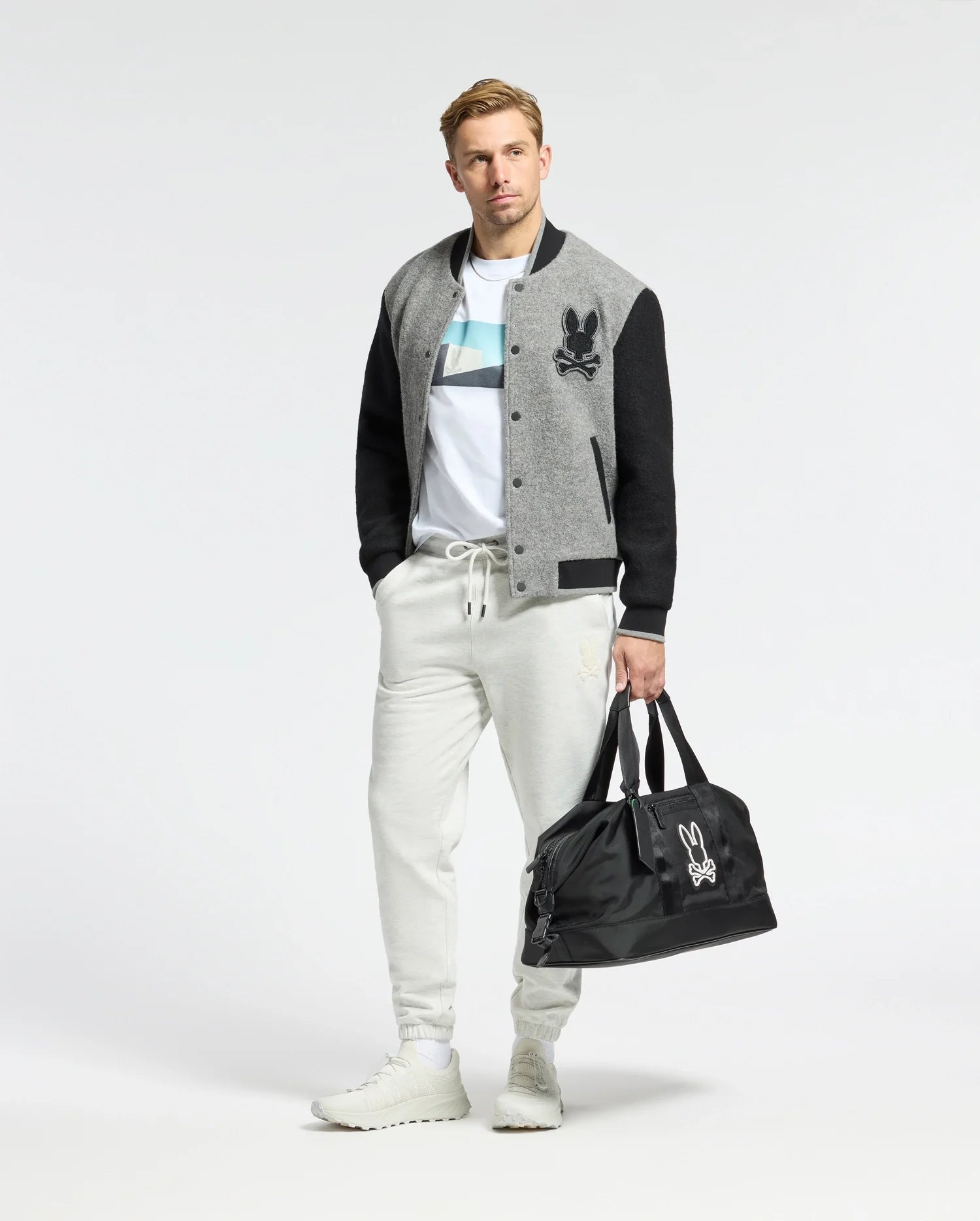 A person stands against a plain background wearing a gray and black varsity jacket, Psycho Bunny MENS NEIL PHOTO REAL TEE in premium Peruvian Pima cotton, light gray joggers, and white sneakers. They're holding a black duffel bag with a white logo in one hand.