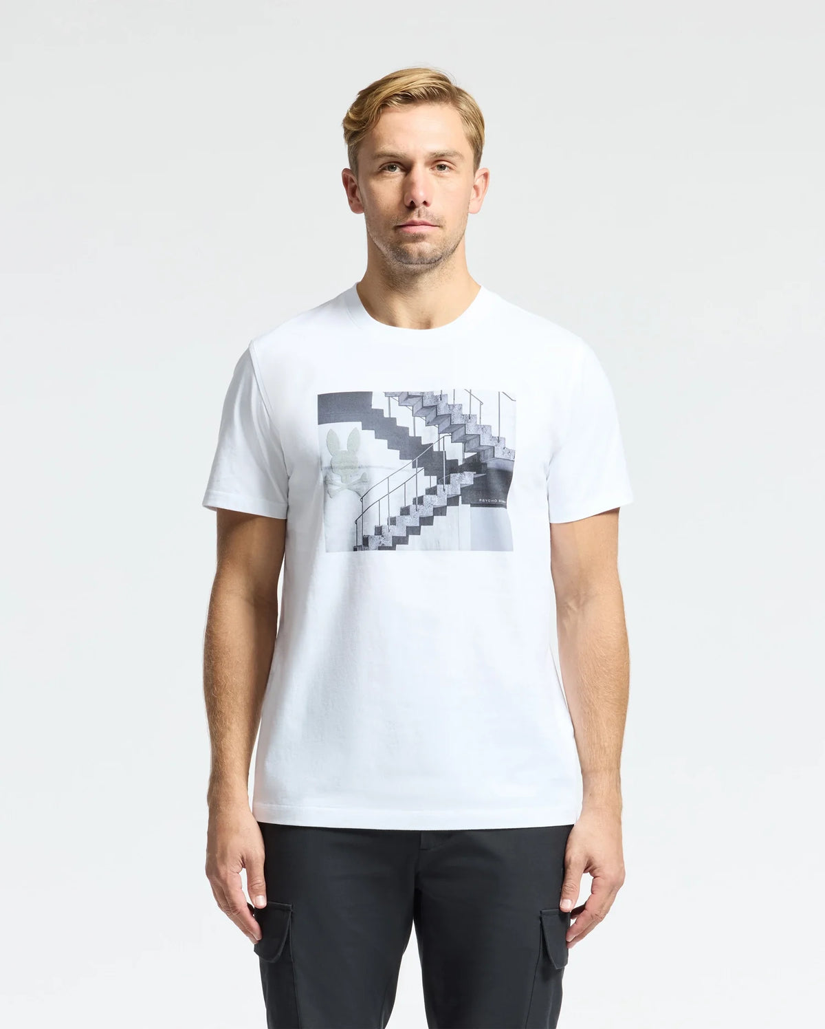 A man in a white Psycho Bunny MENS DEAN PHOTO REAL TEE - B6U390E200, showcasing urban geometric black-and-white staircase designs, stands against a plain backdrop. Made from soft Pima cotton, he has short hair and gazes directly at the camera.