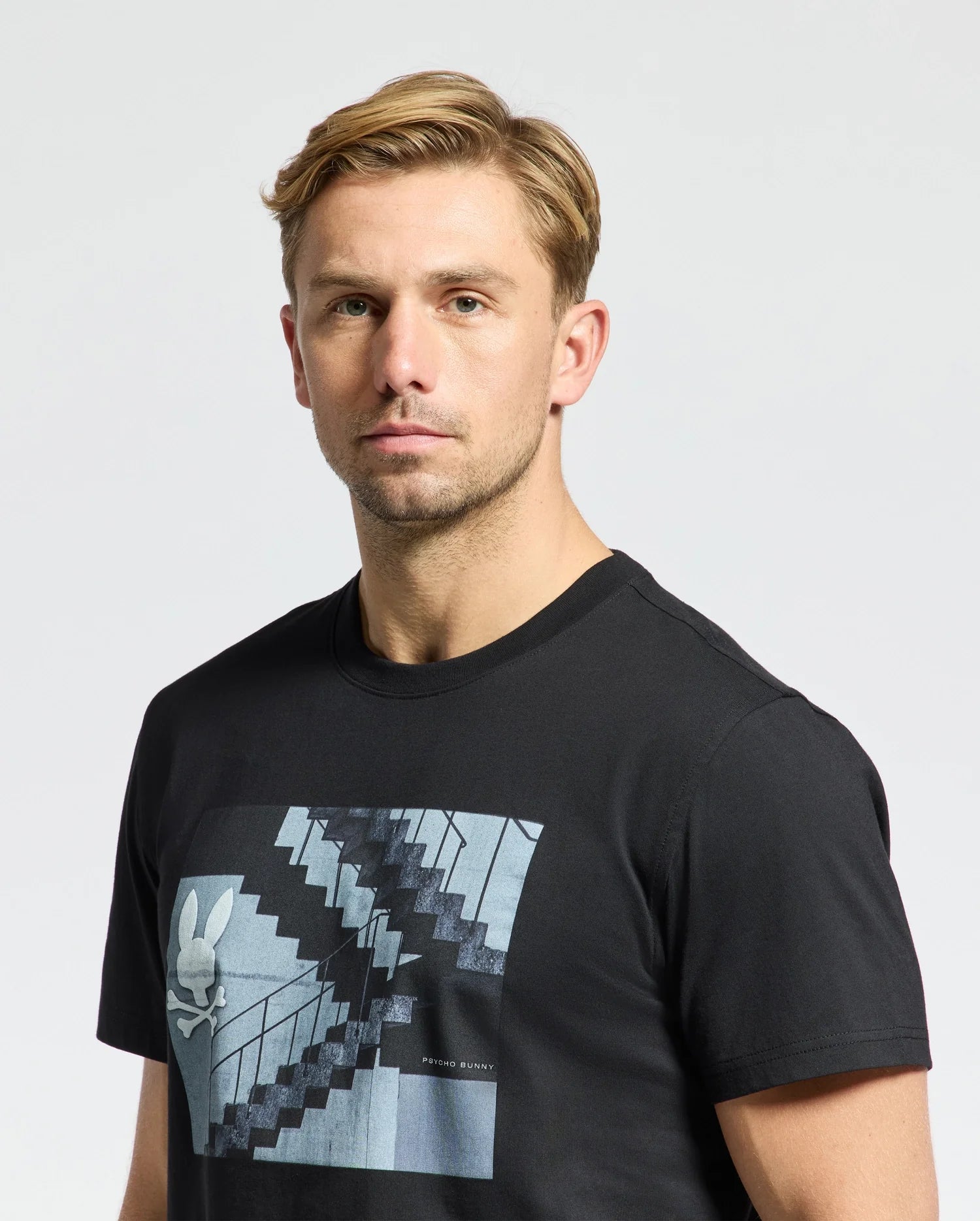 A man with short blond hair wears a stylish Psycho Bunny MENS DEAN PHOTO REAL TEE - B6U390E200, crafted from Pima cotton jersey, featuring a stairs and bunny graphic. The neutral background is light in color.