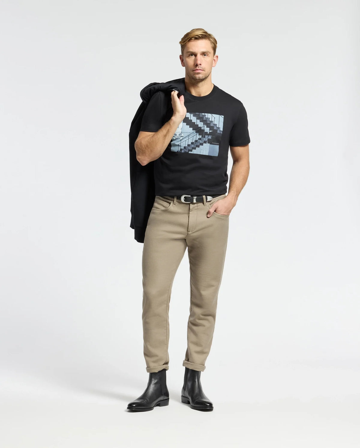 A man confidently wears a Psycho Bunny MENS DEAN PHOTO REAL TEE (B6U390E200), showcasing its urban geometric design. Styled with beige pants and black boots, he casually drapes a jacket over his shoulder, hand in pocket, posing coolly against a plain white background.