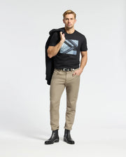 A man confidently wears a Psycho Bunny MENS DEAN PHOTO REAL TEE (B6U390E200), showcasing its urban geometric design. Styled with beige pants and black boots, he casually drapes a jacket over his shoulder, hand in pocket, posing coolly against a plain white background.