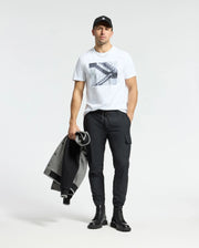 Wearing a Psycho Bunny MENS DEAN PHOTO REAL TEE, black jogger pants, boots, and cap, a man holds a sleek gray and black coat. The minimalistic backdrop enhances his urban style with subtle geometric shapes.