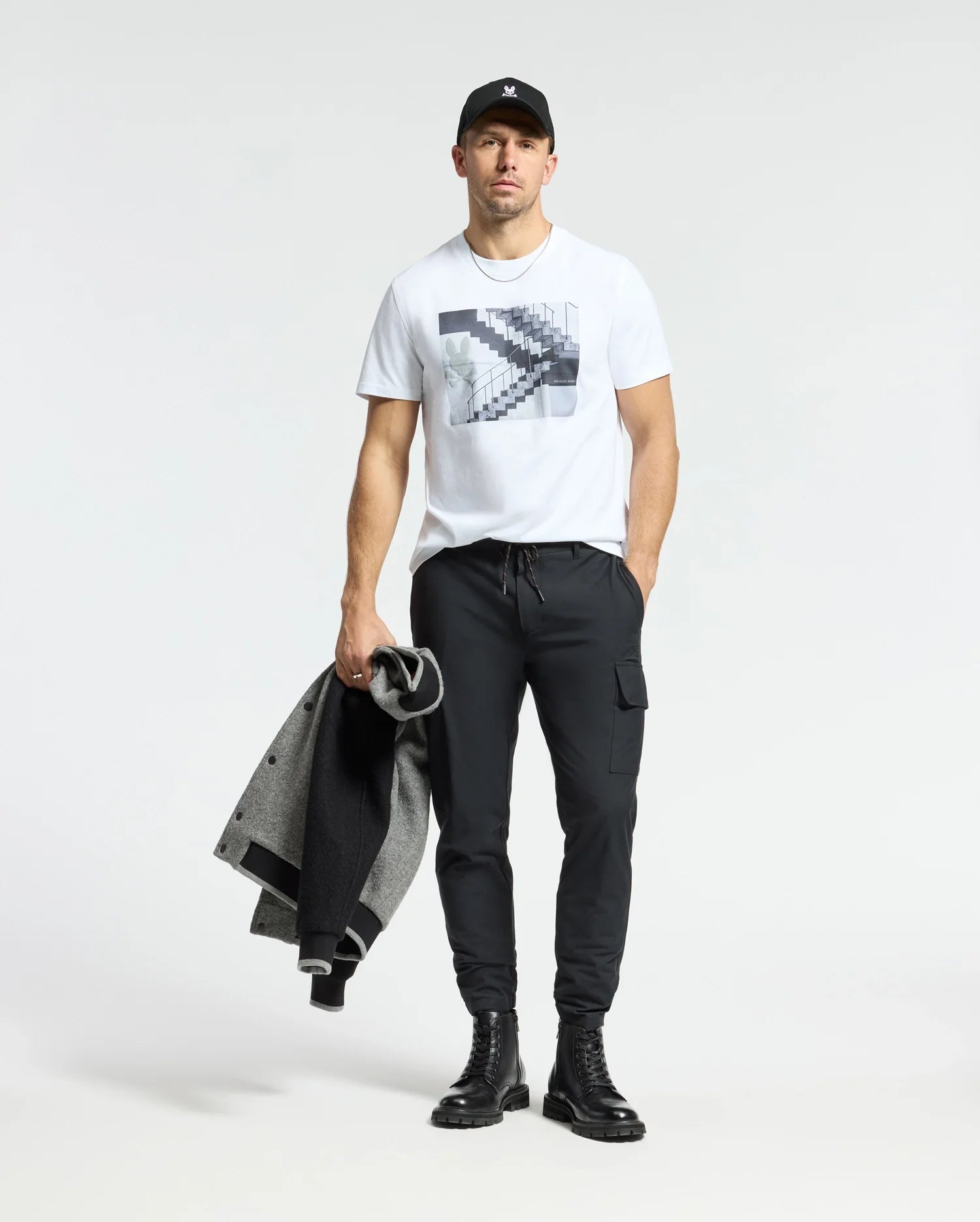 Wearing a Psycho Bunny MENS DEAN PHOTO REAL TEE, black jogger pants, boots, and cap, a man holds a sleek gray and black coat. The minimalistic backdrop enhances his urban style with subtle geometric shapes.