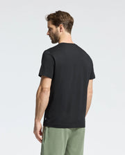 A short-haired, bearded man faces away wearing Psycho Bunny's MENS MELBOURNE GRAPHIC TEE (B6U410F200) in black with a bunny graphic, paired with sage green shorts against a plain white background.
