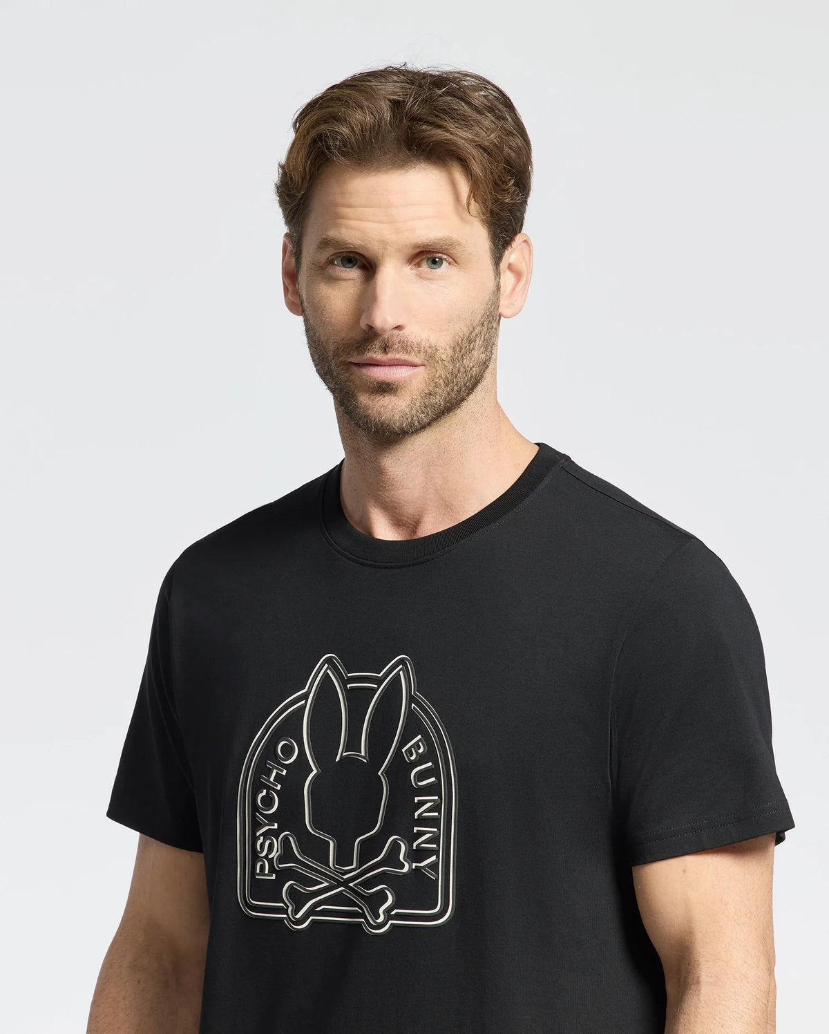 A short brown-haired, bearded man stands against a plain light background, wearing the Psycho Bunny MENS MELBOURNE GRAPHIC TEE - B6U410F200 featuring Peruvian Pima knit with a bold Bunny graphic and crossbones design.