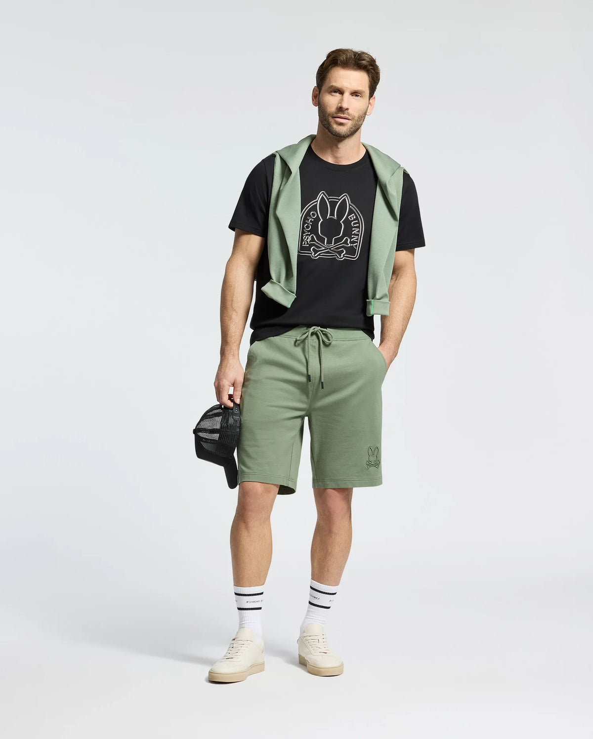 A man stands wearing a Psycho Bunny MENS MELBOURNE GRAPHIC TEE (B6U410F200), light green shorts, and a matching sweatshirt draped over his shoulders. He holds a black cap, wearing white socks with sneakers, against a plain white background.