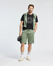 A man stands wearing a Psycho Bunny MENS MELBOURNE GRAPHIC TEE (B6U410F200), light green shorts, and a matching sweatshirt draped over his shoulders. He holds a black cap, wearing white socks with sneakers, against a plain white background.
