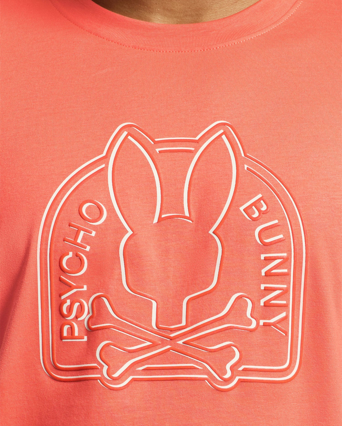 Close-up of a coral MENS MELBOURNE GRAPHIC TEE by Psycho Bunny, made from eco-friendly Peruvian Pima Cotton, featuring an embossed Bunny graphic with elongated ears atop crossed bones and the words "PSYCHO BUNNY" encircling the logo.