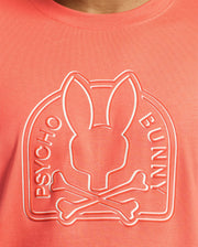 Close-up of a coral MENS MELBOURNE GRAPHIC TEE by Psycho Bunny, made from eco-friendly Peruvian Pima Cotton, featuring an embossed Bunny graphic with elongated ears atop crossed bones and the words "PSYCHO BUNNY" encircling the logo.