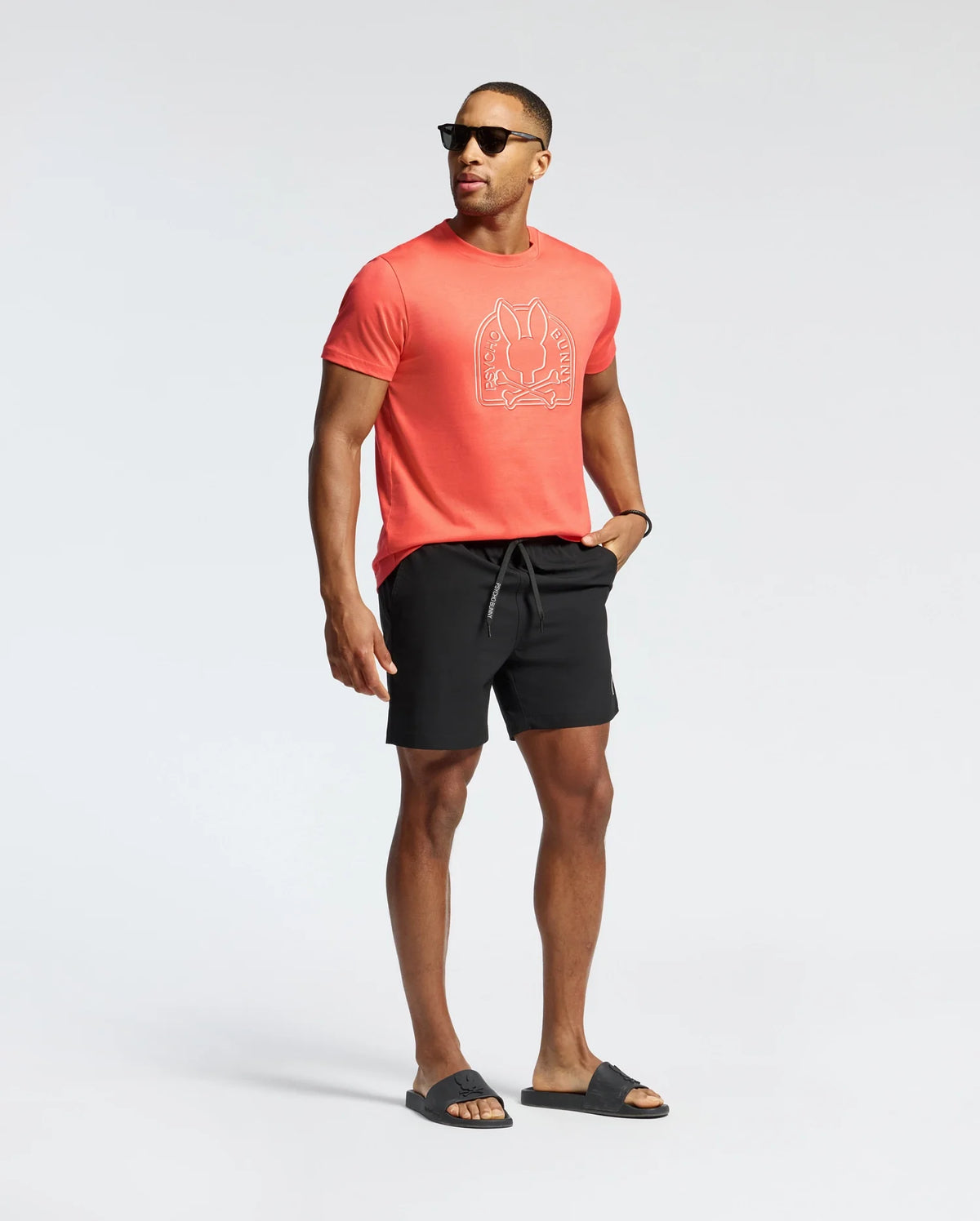 A man wearing sunglasses and a vibrant orange Psycho Bunny MENS MELBOURNE GRAPHIC TEE - B6U410F200, crafted from eco-friendly Peruvian Pima Cotton, confidently stands against a plain background. He's paired it with black shorts and sandals, posing with one hand in his pocket.