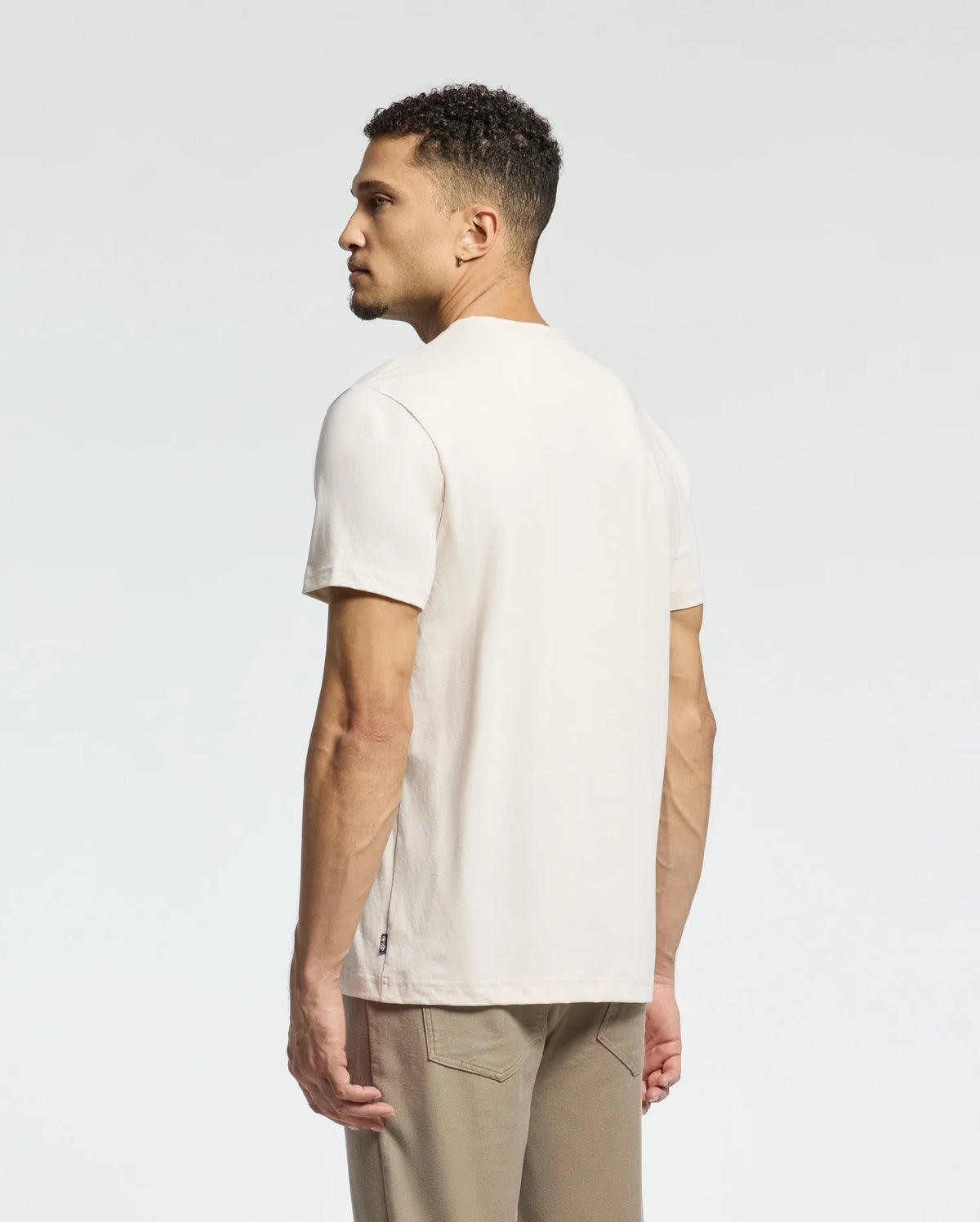 A man with short curly hair faces away, wearing a Psycho Bunny Men's Melbourne Graphic Tee in light beige and light brown pants. Crafted from luxurious Peruvian Pima cotton, this eco-friendly style offers a minimalistic look against a soft white-to-light gray gradient.