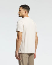 A man with short curly hair faces away, wearing a Psycho Bunny Men's Melbourne Graphic Tee in light beige and light brown pants. Crafted from luxurious Peruvian Pima cotton, this eco-friendly style offers a minimalistic look against a soft white-to-light gray gradient.