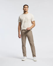 A man wearing a Psycho Bunny MENS MELBOURNE GRAPHIC TEE - B6U410F200, crafted from Peruvian Pima cotton, pairs it with khaki pants and white sneakers. Holding sunglasses in one hand, he shows off an eco-friendly vibe against a plain backdrop.