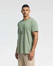 A person in an eco-friendly light green MENS MELBOURNE GRAPHIC TEE (B6U410F200) by Psycho Bunny stands against a white backdrop. They're dressed in beige pants and face forward with a neutral look.