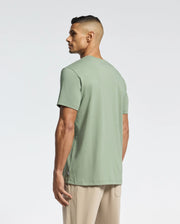 A man with short dark hair models a light green, eco-friendly Psycho Bunny MENS MELBOURNE GRAPHIC TEE - B6U410F200 and beige pants, standing sideways while looking left against a plain white background.
