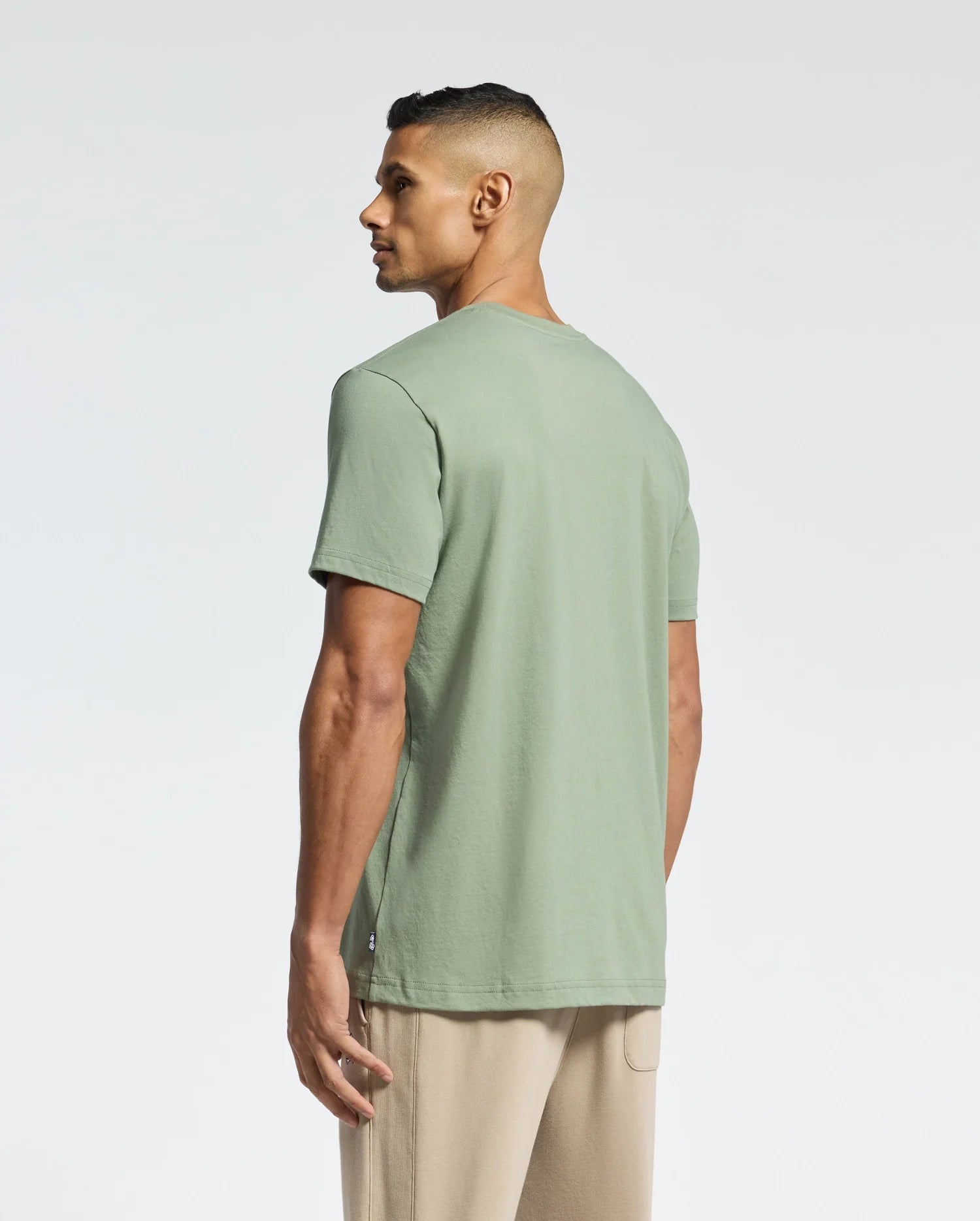 A man with short dark hair models a light green, eco-friendly Psycho Bunny MENS MELBOURNE GRAPHIC TEE - B6U410F200 and beige pants, standing sideways while looking left against a plain white background.