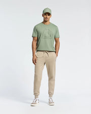 Wearing a green Psycho Bunny MENS MELBOURNE GRAPHIC TEE - B6U410F200 featuring Bugs Bunny, paired with beige joggers and white sneakers, a person stands against a plain white background. This eco-friendly outfit is casual yet stylish, ideal for relaxing or heading out.