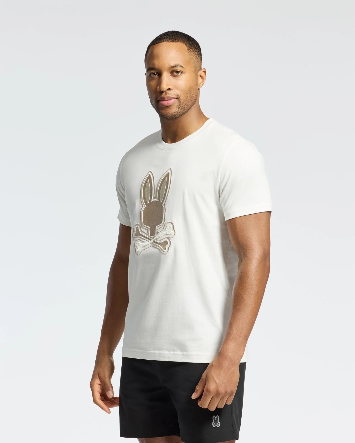 A man stands against a light backdrop wearing a white Psycho Bunny Men's New Castle Graphic Tee - B6U415F200, featuring an embroidered Bunny skull logo and crafted from soft Pima cotton, paired with sleek black shorts. His short hair complements the minimalist style.