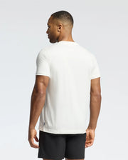 A man wearing a Psycho Bunny MENS NEW CASTLE GRAPHIC TEE and black shorts stands sideways to the camera, his head turned slightly left against a plain white background.