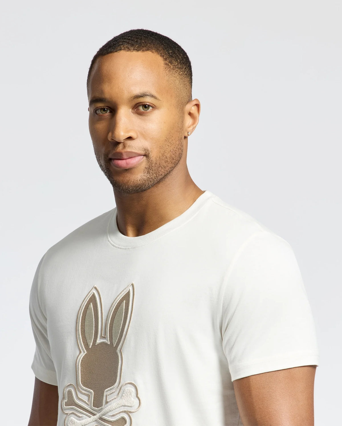 A man stands against a plain backdrop wearing a light-colored Psycho Bunny MENS NEW CASTLE GRAPHIC TEE (B6U415F200) with an embroidered bunny logo.