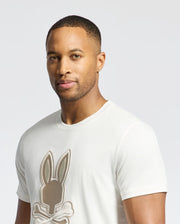 A man stands against a plain backdrop wearing a light-colored Psycho Bunny MENS NEW CASTLE GRAPHIC TEE (B6U415F200) with an embroidered bunny logo.