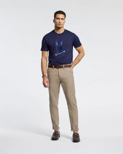 A man wears the Psycho Bunny MENS NEW CASTLE GRAPHIC TEE - B6U415F200, a navy blue Pima cotton tee with an embroidered bunny design, against a white background. He completes his look with beige pants, brown leather shoes, and a matching belt.