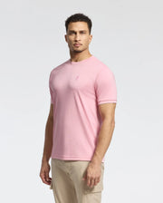 A man with short curly hair wears the Psycho Bunny MENS BRYCE PERFORMANCE TEE in pink, made from a premium Modal-Pima cotton blend, featuring a minimalist logo. Paired with beige pants, the Peruvian-made outfit stands out against a plain white background.