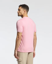 A man with curly hair stands leftward in a pink Psycho Bunny MENS BRYCE PERFORMANCE TEE - B6U490F200, made from a Modal-Pima cotton blend. Paired with beige pants, he poses against a plain white background. This stylish Peruvian-made piece offers both comfort and quality.