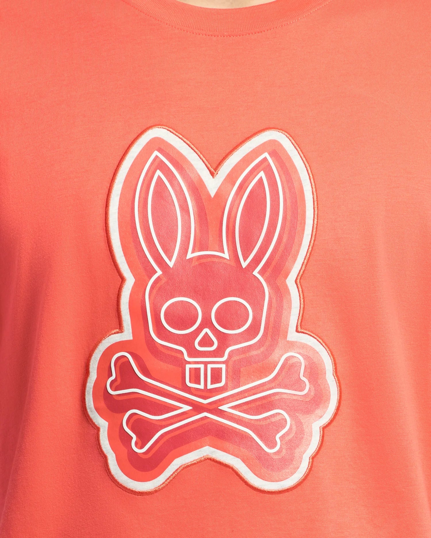 The Mens Sydney Graphic Tee by Psycho Bunny is a coral-colored T-shirt featuring a stylized graphic of a bunny with a skull face and crossbones. Made from soft Pima cotton, it displays layered pink shades with a white outline, offering an edgy twist on classic prints.