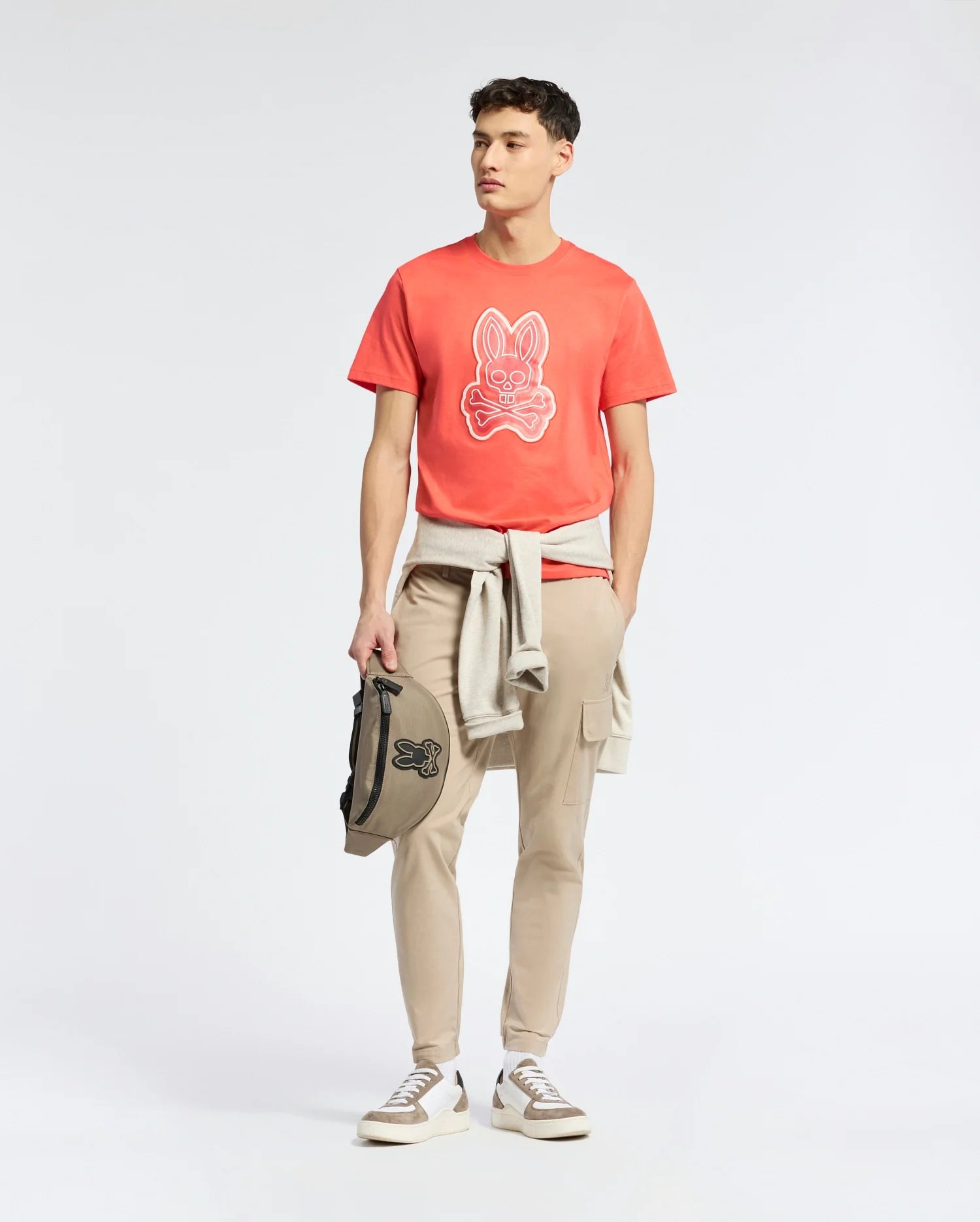 A man casually dons the Psycho Bunny MENS SYDNEY GRAPHIC TEE - B6U508F200 with an animal face print, teamed with beige joggers and white sneakers. A gray sweatshirt is tied around his waist while he carries a black bag, all set against a plain white backdrop.
