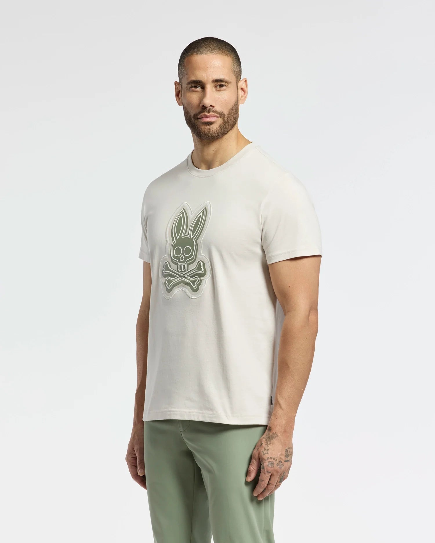 A person with short dark hair and a beard sports the Psycho Bunny MENS SYDNEY GRAPHIC TEE (B6U508F200), a cream Pima cotton t-shirt featuring a bunny skull and crossed bones graphic, paired with light green pants against a plain white background.