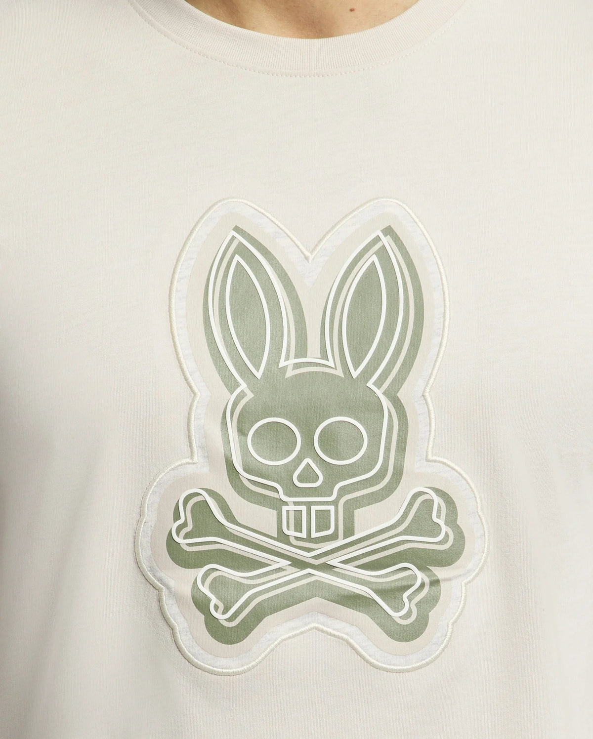 The Psycho Bunny MENS SYDNEY GRAPHIC TEE (B6U508F200) features a bold white bunny graphic with long ears blending into a skull over crossbones on a muted green background. Made from soft Pima cotton, it exemplifies unique and striking designs.
