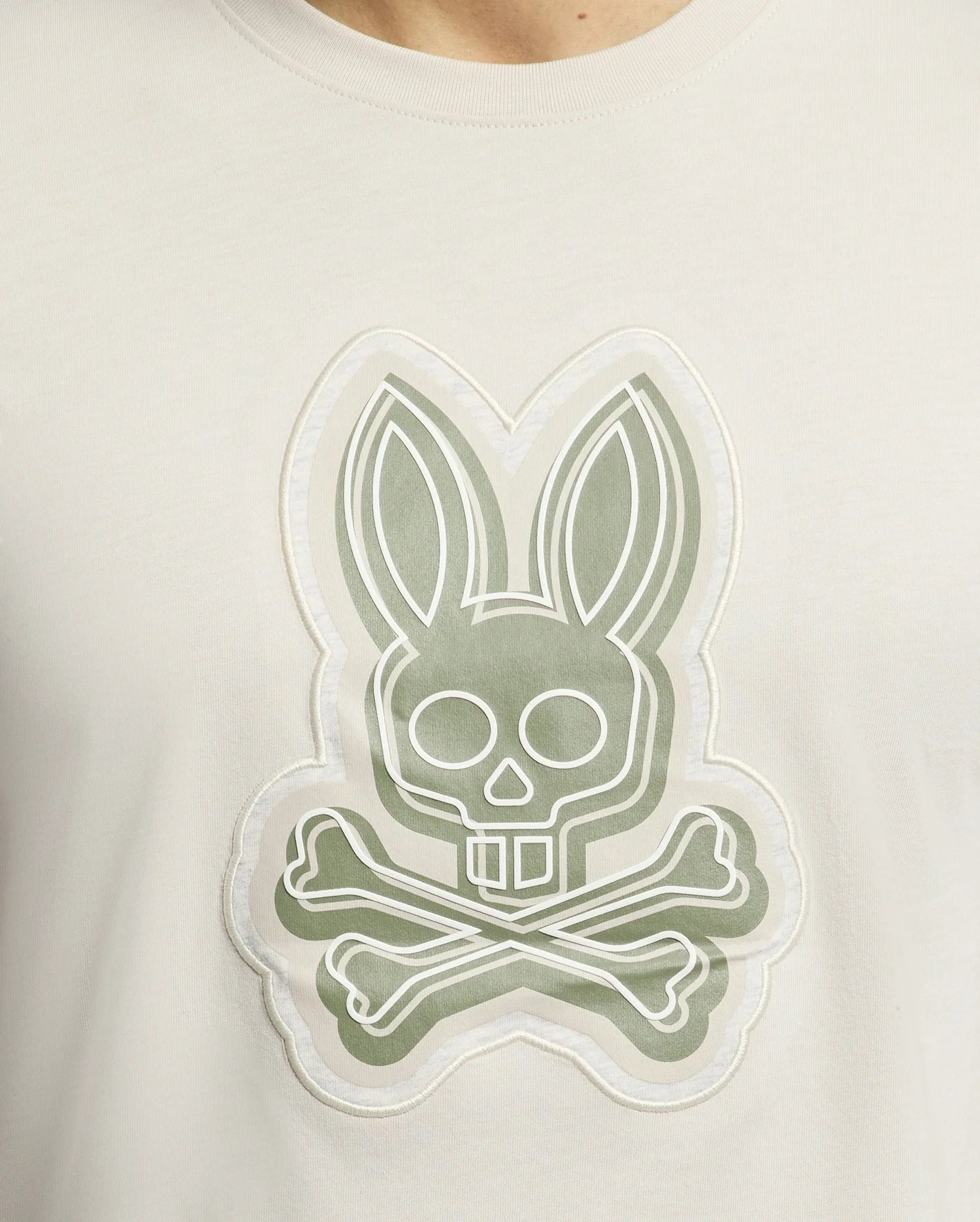 The Psycho Bunny MENS SYDNEY GRAPHIC TEE (B6U508F200) features a bold white bunny graphic with long ears blending into a skull over crossbones on a muted green background. Made from soft Pima cotton, it exemplifies unique and striking designs.