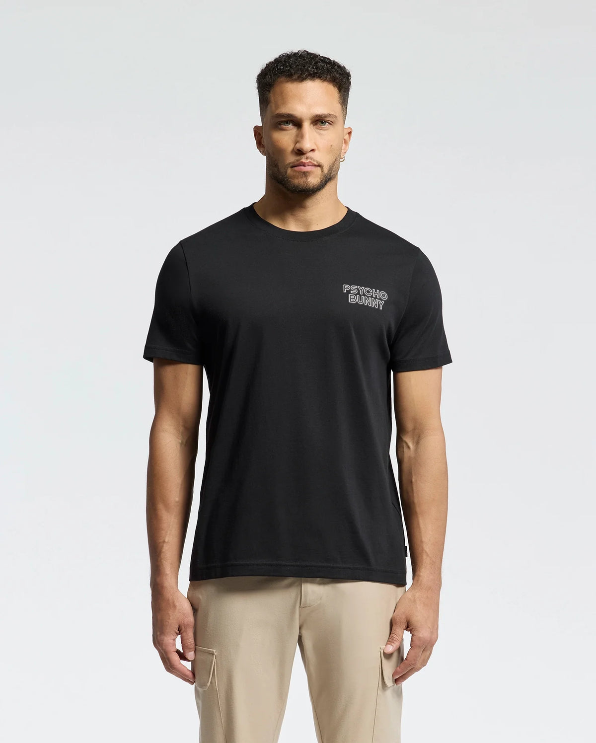A man in a regular fit Psycho Bunny MENS SYDNEY BACK GRAPHIC TEE, crafted from soft Pima cotton with a small chest print, stands against a plain background. His short curly hair and beard complement his casual look, completed with beige pants.