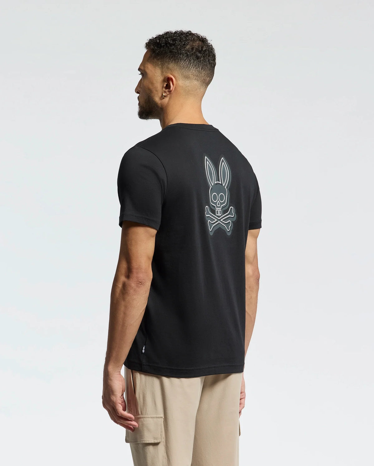 A man with short curly hair, back to the camera, wears a Psycho Bunny MENS SYDNEY BACK GRAPHIC TEE - B6U509F200 in regular fit black Pima cotton with a water-based bunny skull design. He pairs it with beige cargo pants against a plain, light-colored background.