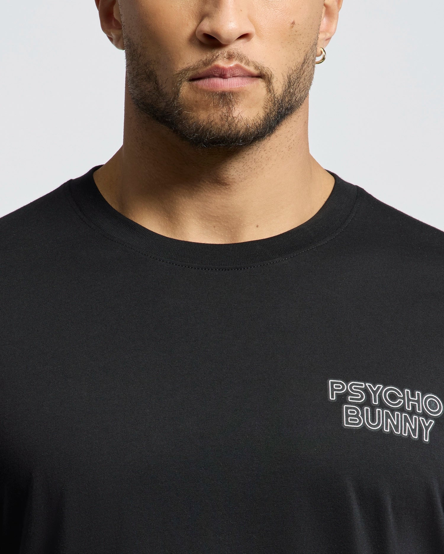 A person wears a MENS SYDNEY BACK GRAPHIC TEE - B6U509F200, crafted from soft Pima cotton in black with a regular fit and short sleeves. The white water-based "Psycho Bunny" print appears on the chest. The person's face is partially visible, cropped at the forehead.