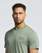 A man in a light green "MENS SYDNEY BACK GRAPHIC TEE - B6U509F200" by Psycho Bunny, crafted from soft Pima cotton, with short curly hair and an earring, looks directly at the camera against a plain backdrop.
