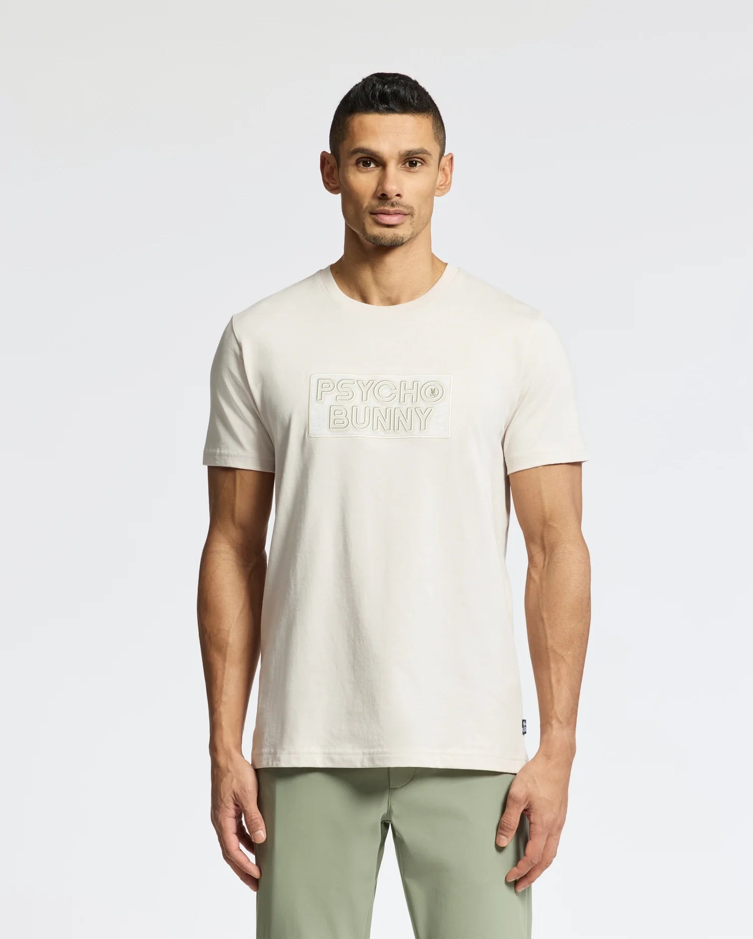 A man in a light-colored Psycho Bunny MENS SYDNEY WORDMARK GRAPHIC TEE (B6U510F200) stands against a plain background. His short, dark hair complements his outfit, which includes light green pants.