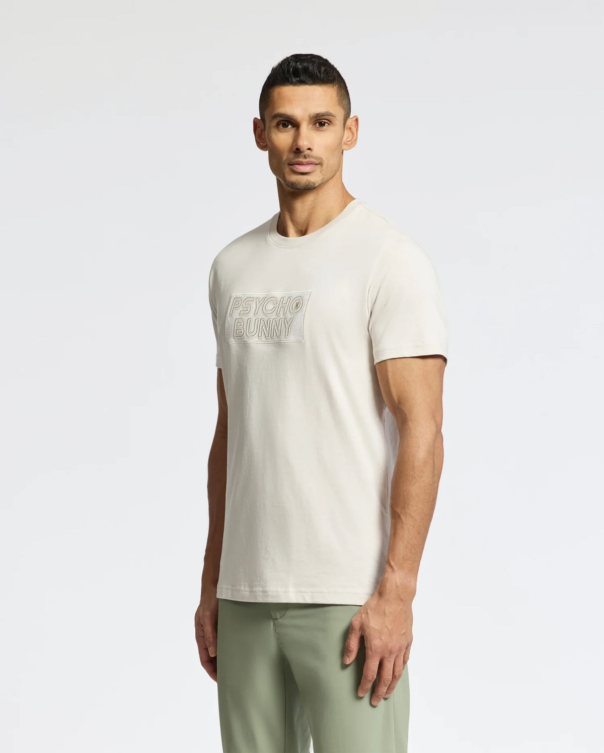A man with short dark hair, against a plain backdrop, sports the MENS SYDNEY WORDMARK GRAPHIC TEE - B6U510F200 by Psycho Bunny, a light beige Pima cotton T-shirt. Paired with light green pants, he achieves a relaxed yet stylish look.