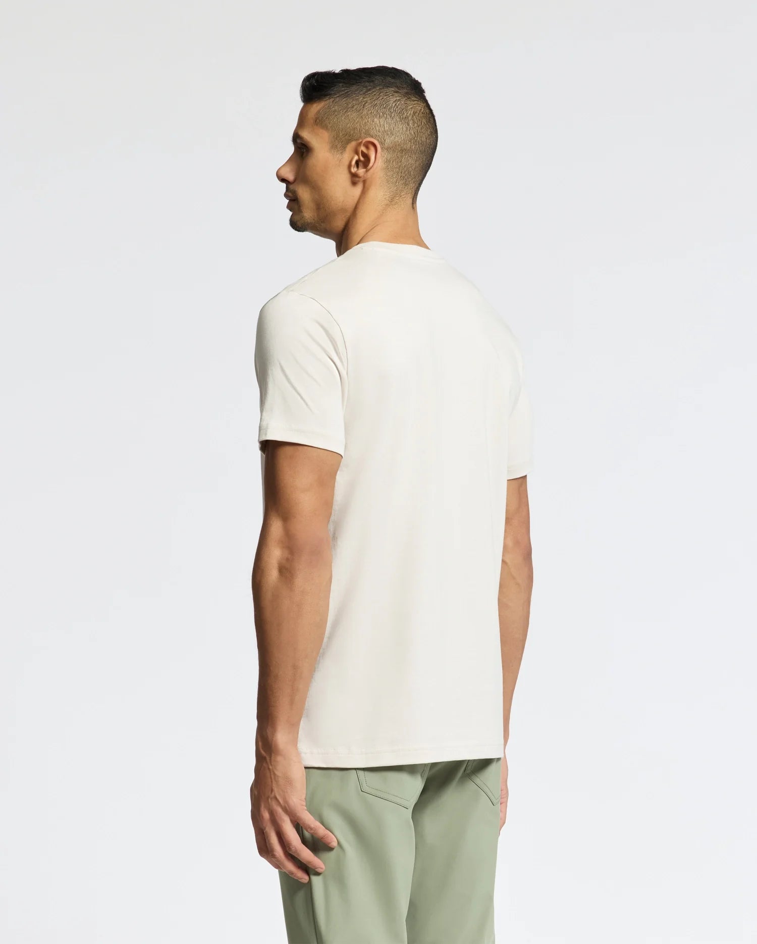 A man, stylishly clad in the Psycho Bunny MENS SYDNEY WORDMARK GRAPHIC TEE - B6U510F200, faces away. The light beige Pima cotton tee features an eco-friendly water-based print and pairs perfectly with olive green pants against a plain white background.