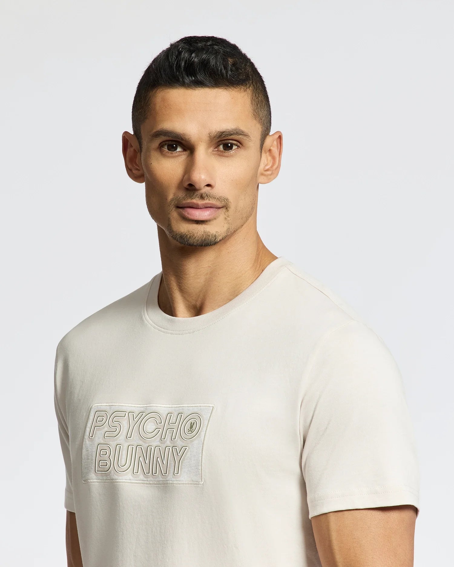 A man with short hair and a trimmed beard, looking serious, wears a luxurious light-colored MENS SYDNEY WORDMARK GRAPHIC TEE - B6U510F200 by Psycho Bunny. The plain, light background enhances his poised demeanor.