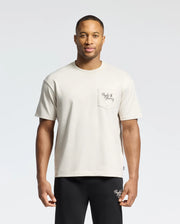 A man wears the Psycho Bunny MENS DESERT OVERSIZED POCKET TEE - B6U512F200, a beige heavyweight jersey T-shirt made from Pima cotton. It has a small text-designed pocket and is paired with black pants. He stands against a plain background with a neutral expression, highlighting its relaxed fit.