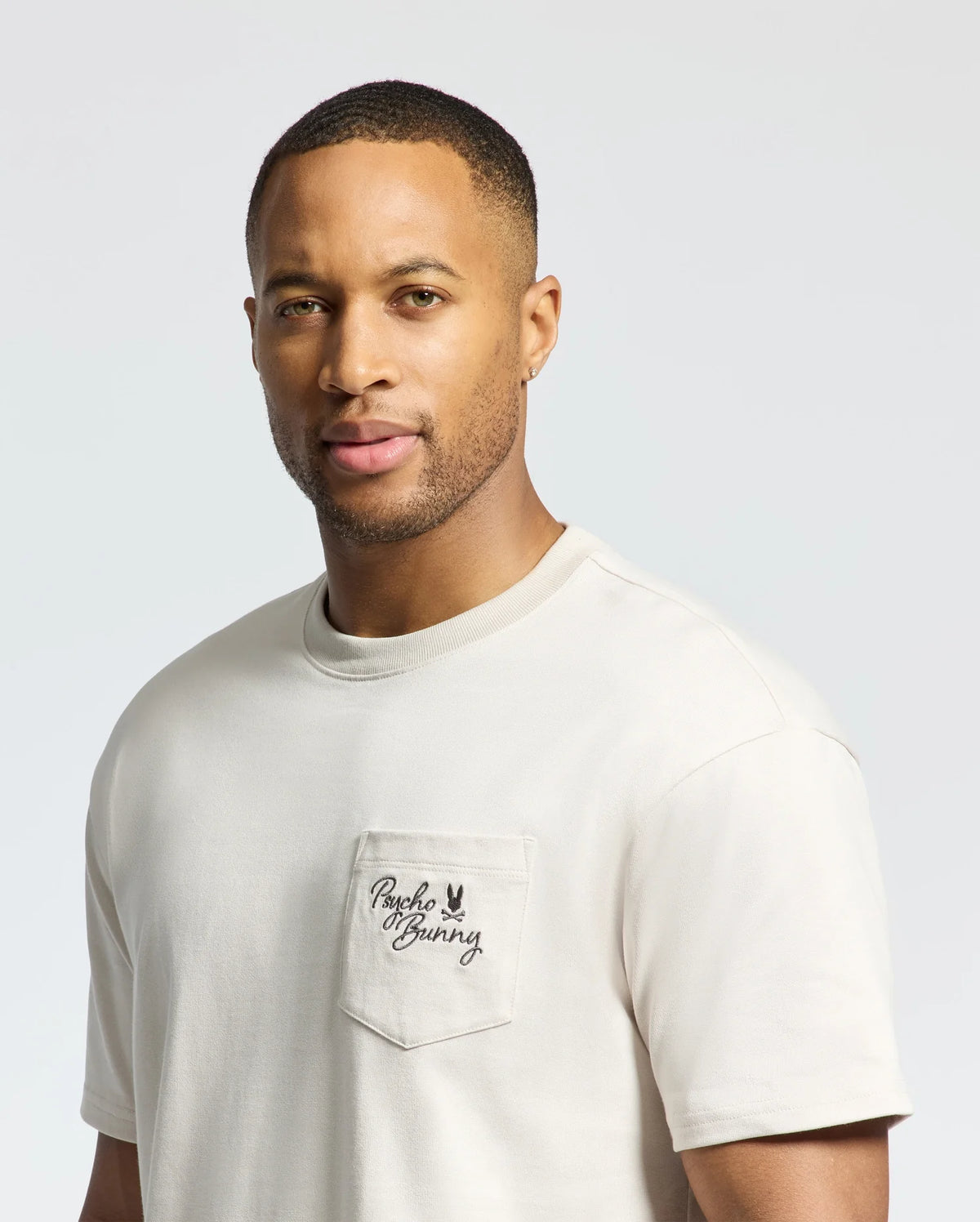 A man in a light beige MENS DESERT OVERSIZED POCKET TEE by Psycho Bunny, crafted from soft Pima cotton with "Psycho Bunny" script on the chest pocket. He has short hair and a neutral expression, with the relaxed fit of the tee enhancing the light gray backdrop.