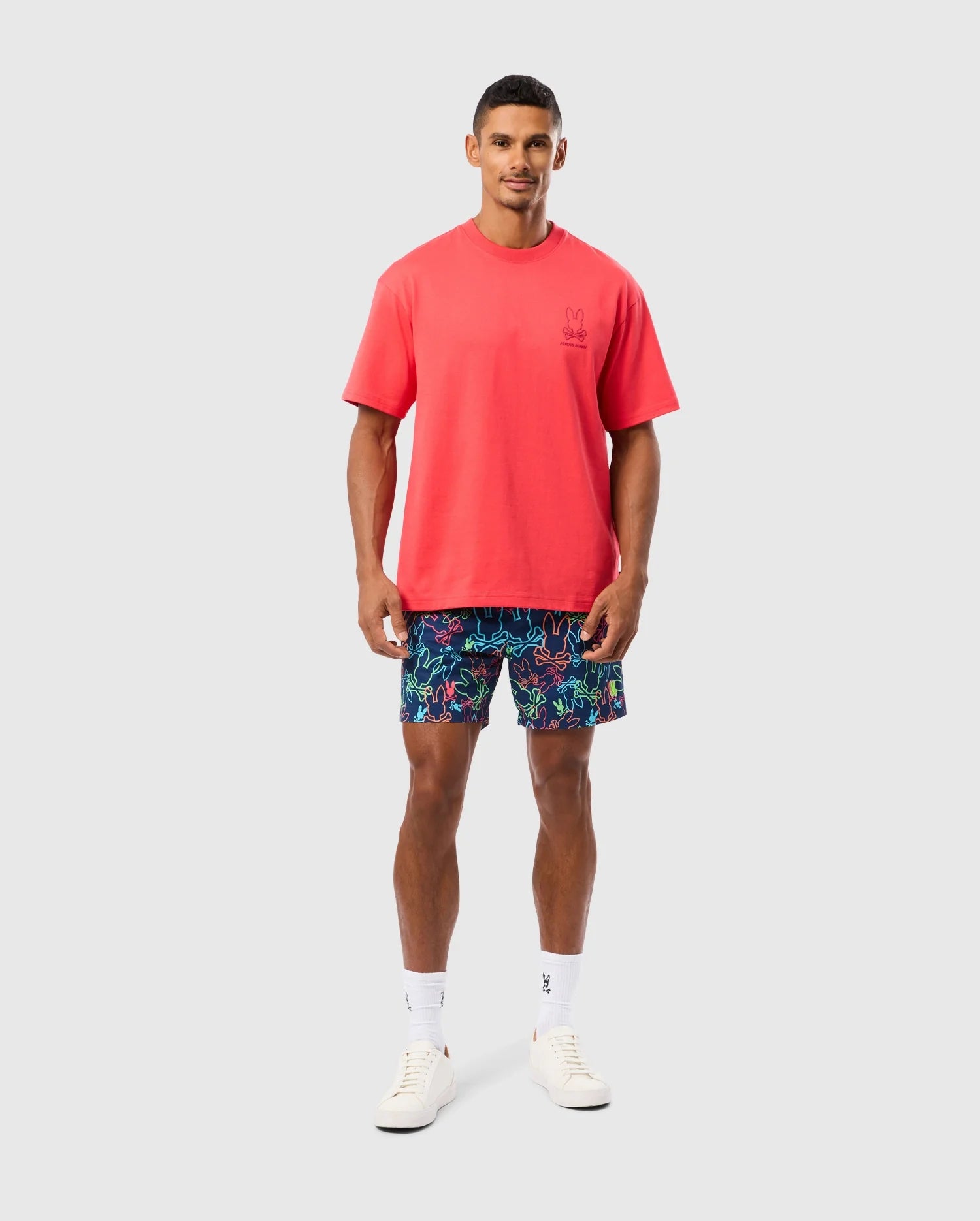 A person with short hair is wearing a bright red Psycho Bunny MENS BARRETT RELAXED FIT TEE - B6U518C200 featuring a Bunny graphic, multicolored shorts with a vibrant pattern, white socks, and white sneakers. The relaxed fit ensures comfort against the plain, light-colored background.