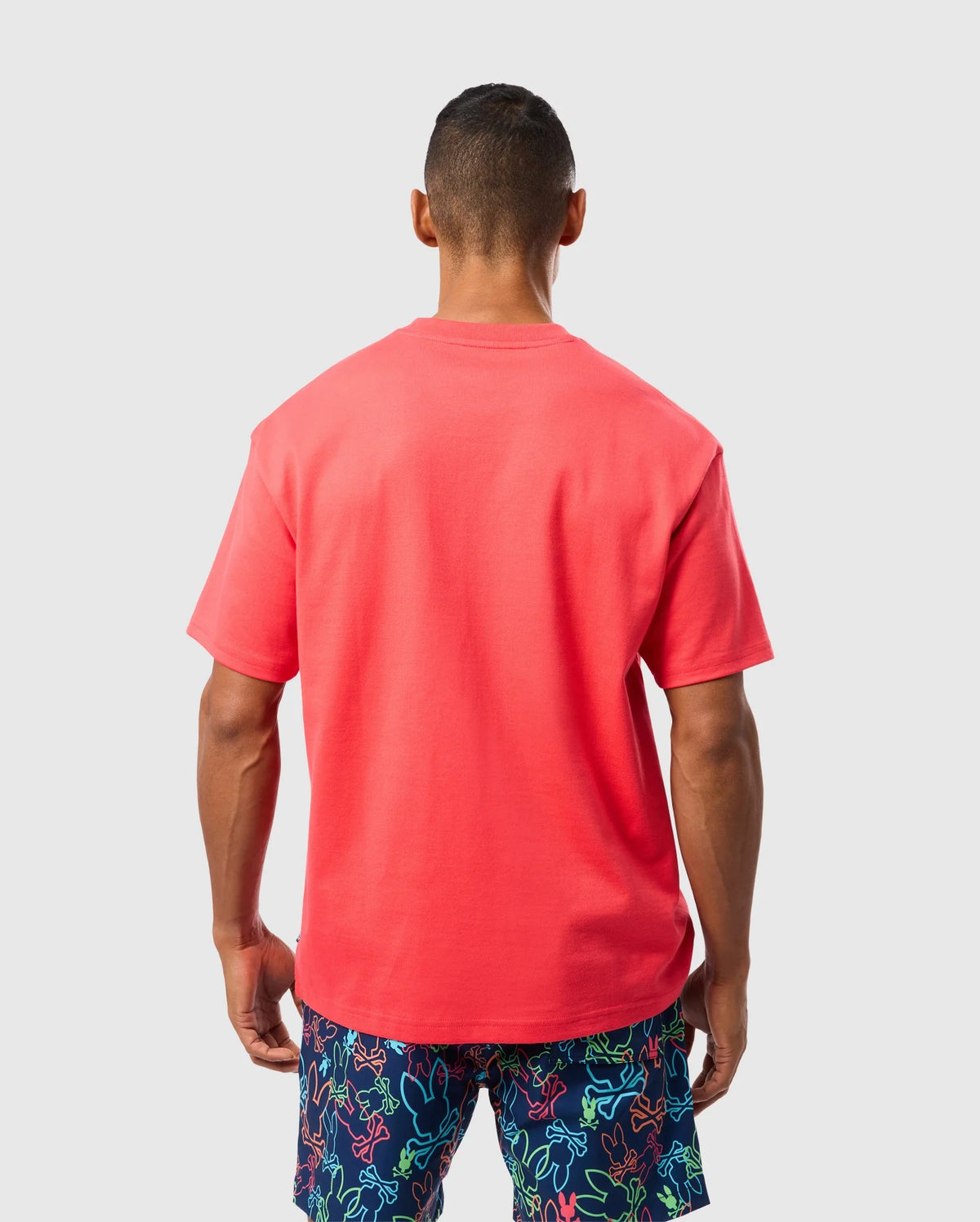 A person with short hair stands facing away, wearing a bright red Psycho Bunny MENS BARRETT RELAXED FIT TEE - B6U518C200 adorned with a subtle bunny graphic and colorful shorts featuring a pattern of various shapes and colors on a light grey background.