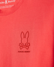 Close-up of a red Psycho Bunny MENS BARRETT RELAXED FIT TEE - B6U518C200 featuring an embossed Bunny graphic with a bunny head and crossbones below it. The text "PSYCHO BUNNY" is stitched under the logo. Made from smooth, heavyweight Pima cotton, the fabric ensures durability while maintaining a relaxed fit.