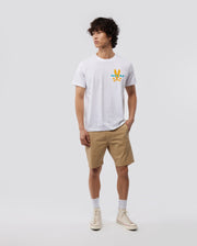 A person with short, curly dark hair is standing against a plain light gray background. They are wearing a Psycho Bunny MENS MIAMI TEE - B6U550W1PC made of soft Pima cotton, beige shorts, white socks, and off-white sneakers. The colorful graphic on the chest adds a vibrant Miami flair as they look slightly to the side.