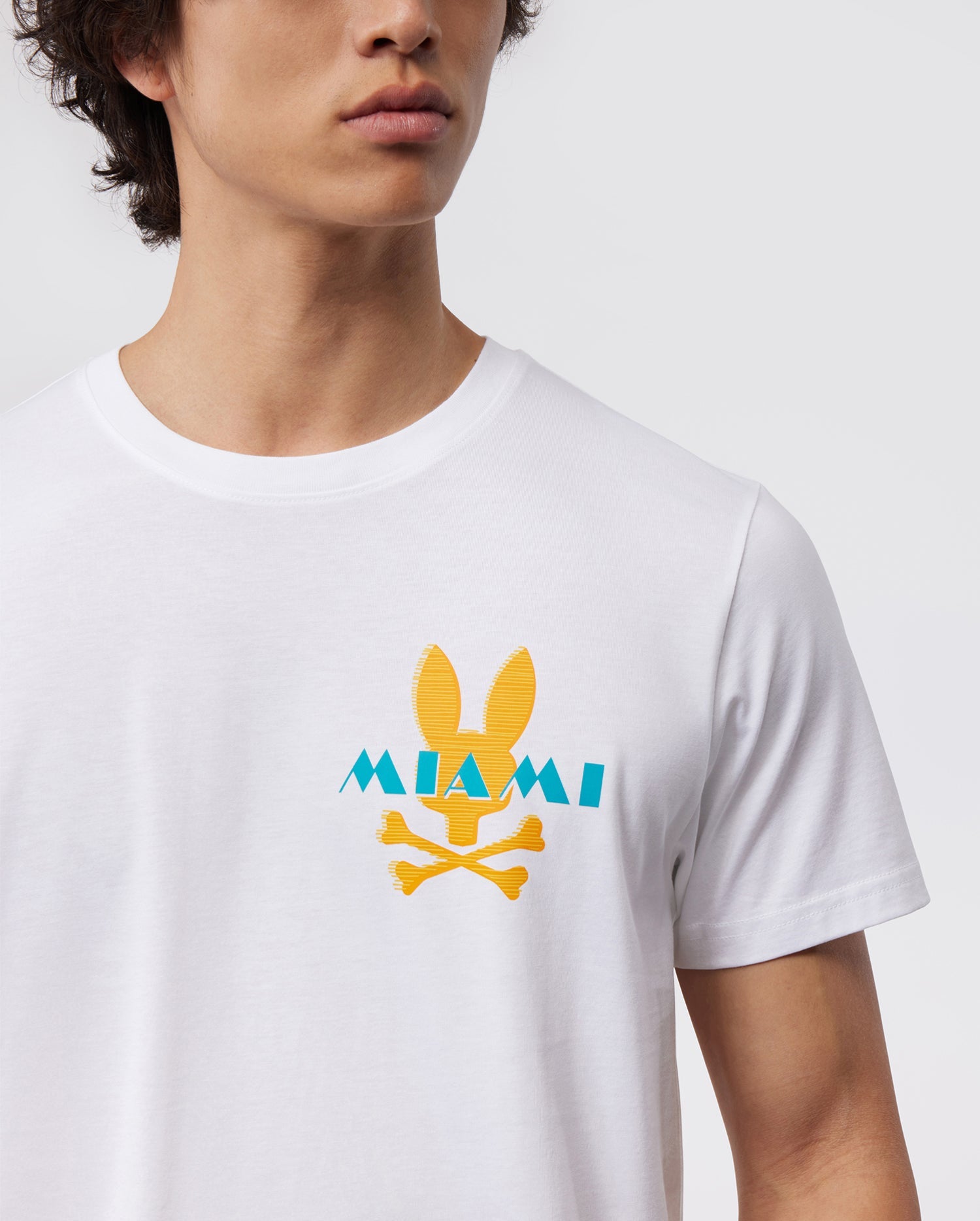 A person wearing a white Pima cotton jersey T-shirt with an orange bunny logo above two crossed bones and the word "MIAMI" written in blue. The person's head is slightly turned to the side, and the background is plain white. This Psycho Bunny MENS MIAMI TEE - B6U550W1PC perfectly embodies casual Miami style.