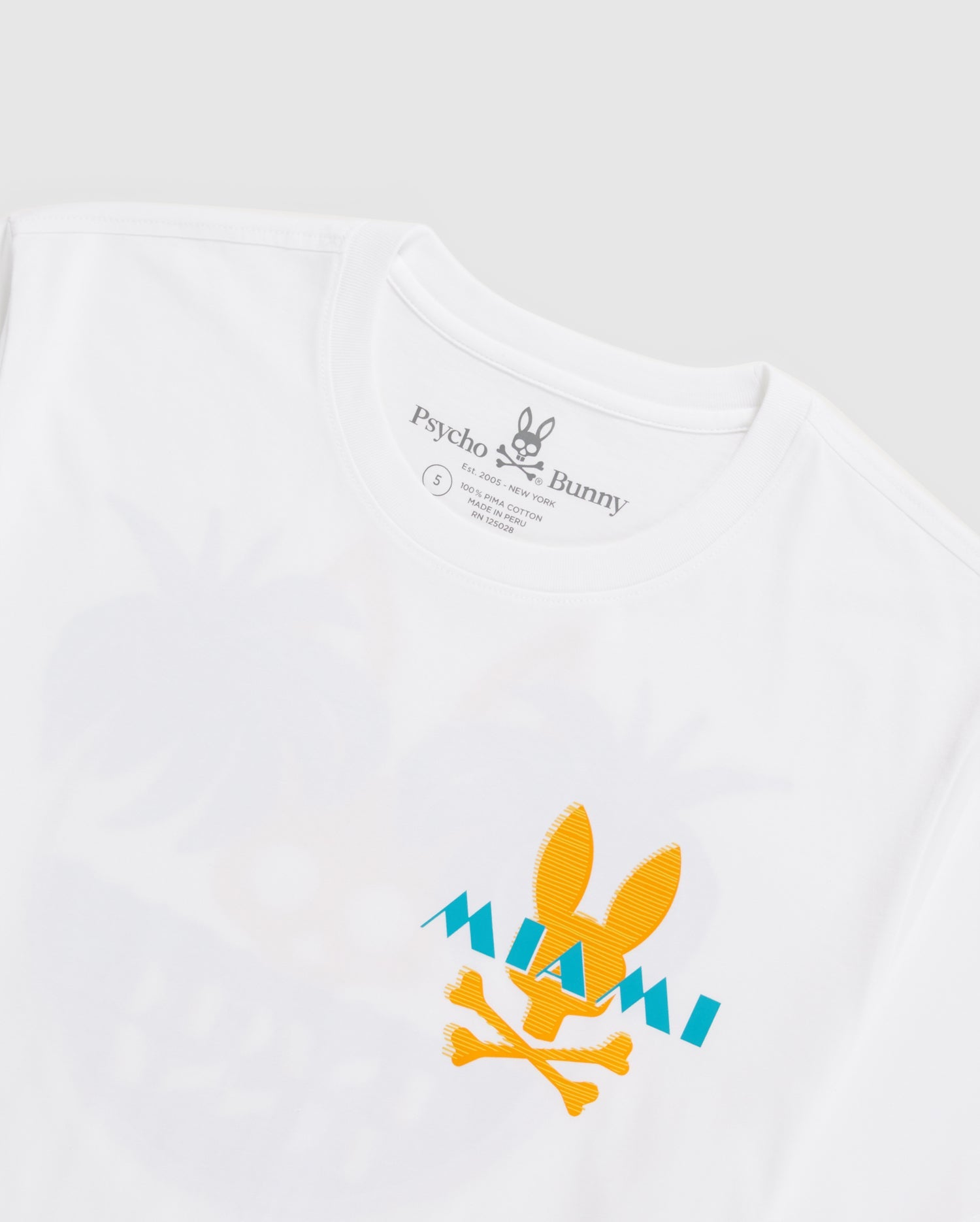 A white men's MENS MIAMI TEE - B6U550W1PC featuring a yellow bunny skull and crossbones with "MIAMI" written in turquoise over it, crafted from soft Pima cotton. The shirt's label reads "Psycho Bunny." The background is a light gray.