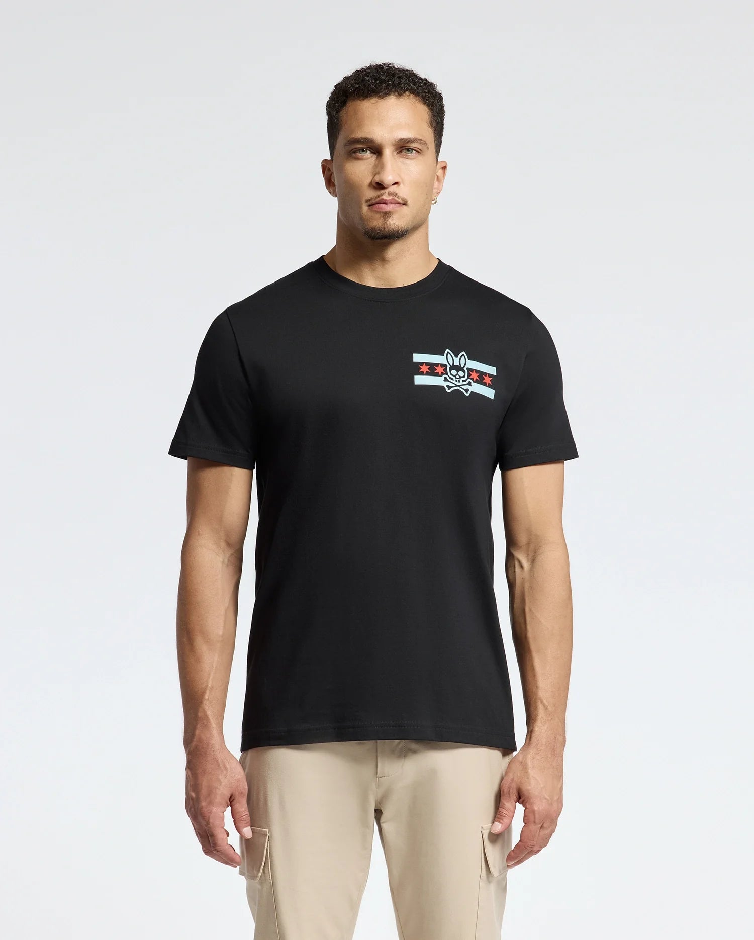 A man wearing the Psycho Bunny MENS CHICAGO TEE - B6U551W1PC, crafted from soft Pima cotton and featuring a small graphic design with stripes and a rabbit emblem on the left chest, is complemented by light beige pants as he stands confidently against a plain white background.